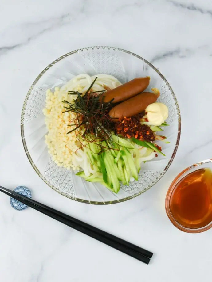 A bowl of cold udon topping with sausages, cucumber, seaweed, tenkasu, with mayonnaise and S&B Chili Oil with Crunchy Garlic