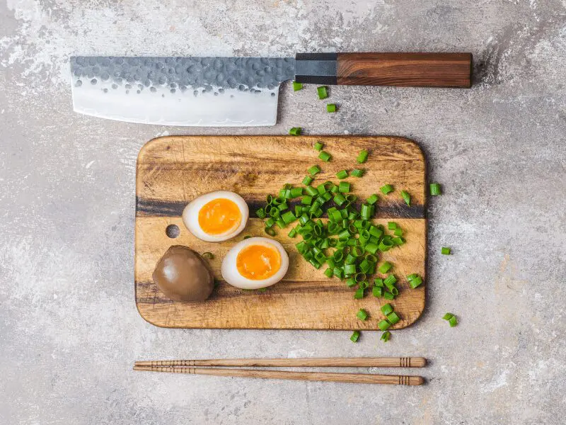 A cutting board with chopped scallions and sliced ajitama on it, a knife and chopsticks parallel to each other.