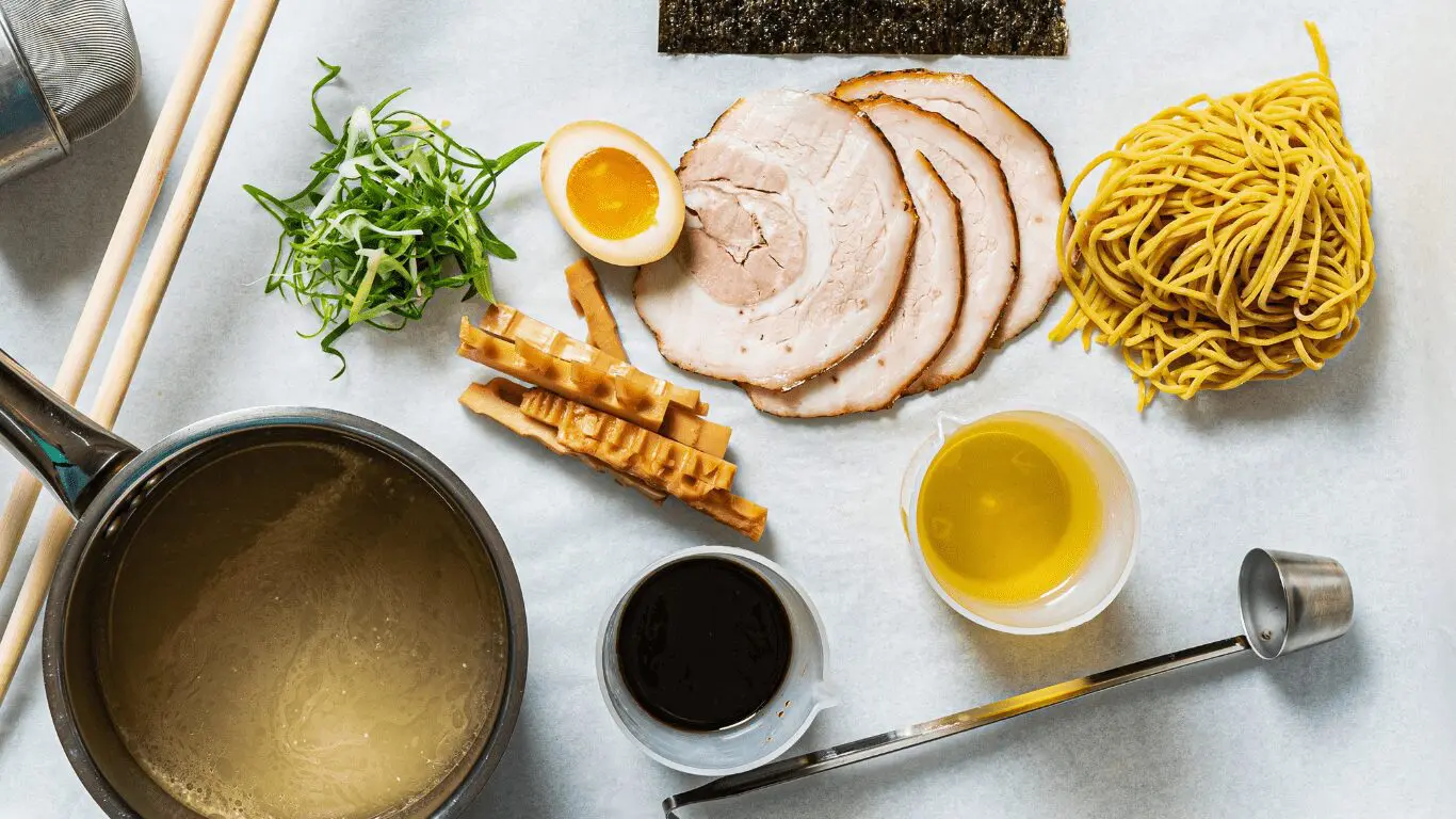 Items related to ramen cooking laid out on the table. From left to right: strainer, chopsticks, soup in a pot, chopped green onions, menma, boiled egg, soy sauce, chashu pork, nori, seasoning oil, noodles, and ladle.