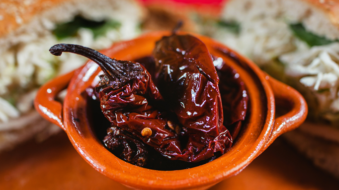 Close-up of chipotle peppers in a small brown clay dish, with blurred sandwiches in the background