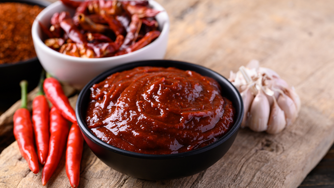 A black bowl filled with gochujang, a thick Korean red chili paste, placed on a wooden surface with fresh red chili peppers, a garlic bulb, and a bowl of dried chilies in the background.