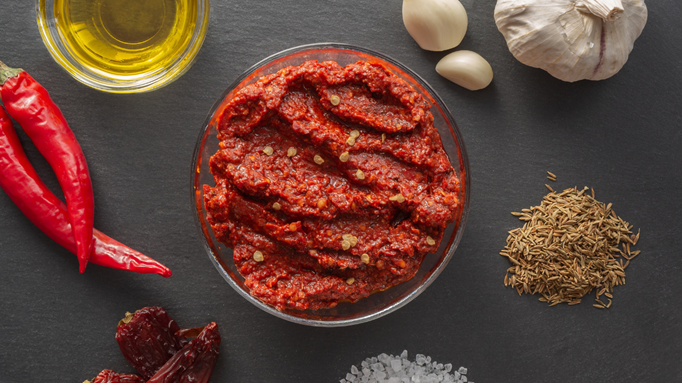 Top view of a bowl of harissa paste surrounded by chili peppers, garlic cloves, cumin seeds, olive oil, and salt on a dark surface