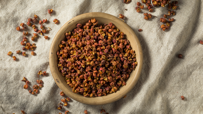 A wooden bowl filled with dried Szechuan peppercorns on a beige cloth surface, with some peppercorns scattered around the bowl