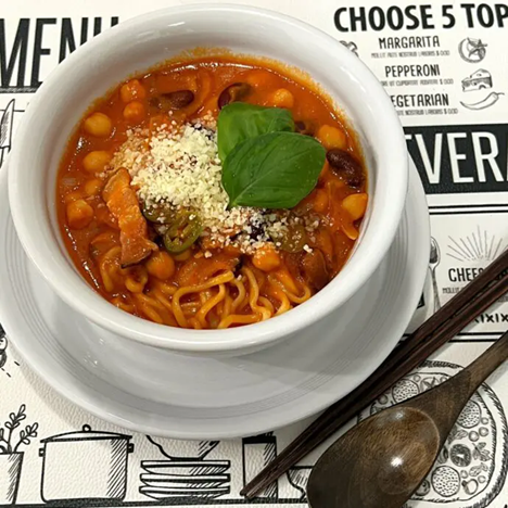 Bowl of chili tomato ramen with beans and vegetables, topped with grated cheese and basil leaf, served on a white plate with chopsticks and spoon.