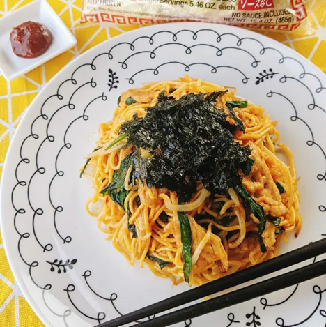 Plate of spicy stir-fried noodles mixed with vegetables and topped with shredded seaweed, served with chopsticks and a small dish of Gochujang paste on the side