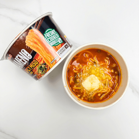 Spicy miso ramen cup shown opened and served, with corn and melted butter as toppings in a rich red broth, next to its original packaging labeled 'Freshly Cooked Noodles