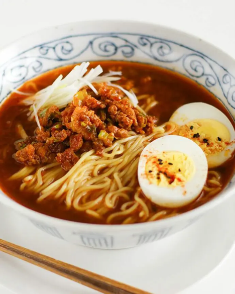 A bowl of spicy miso ramen topped with minced meat, sliced boiled egg, and shredded white scallions in a rich red broth, served in a traditional blue-patterned ceramic bowl