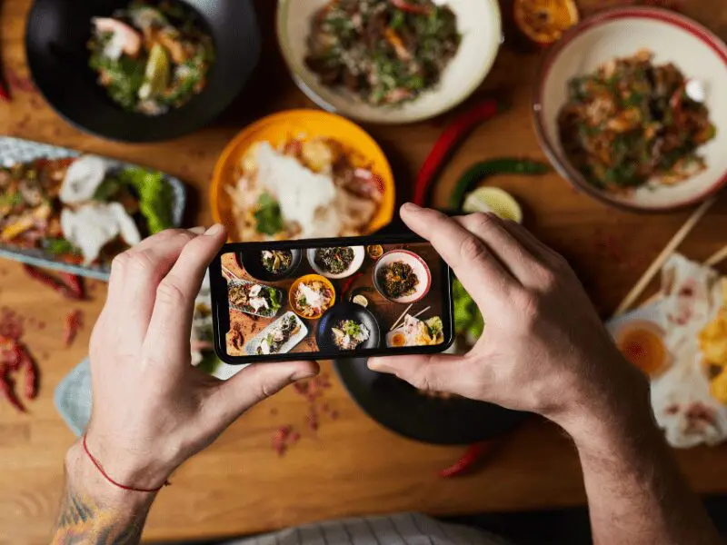 スマートフォンでテーブル上の料理を撮影する人。