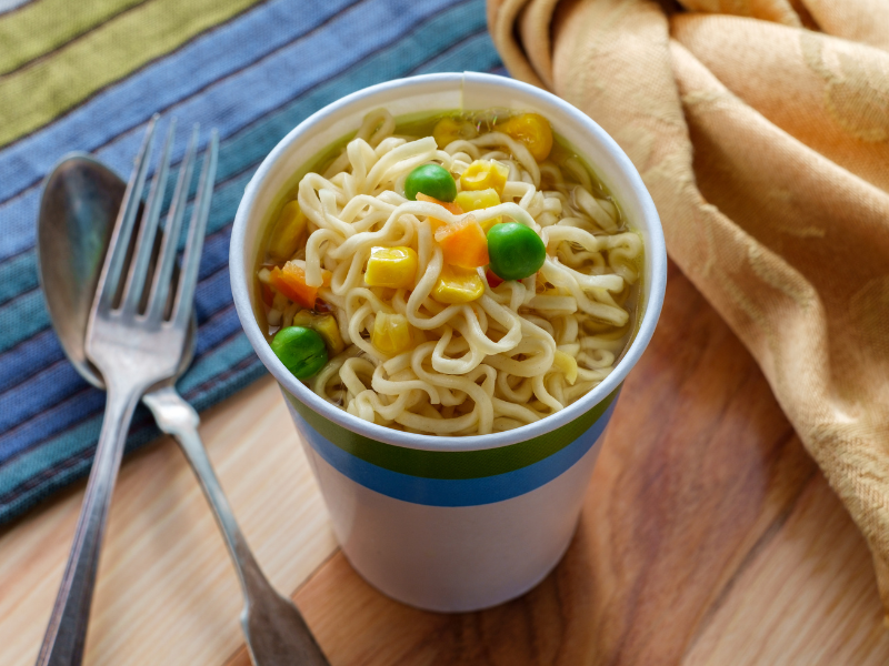 A cup of instant ramen noodles topped with corn, carrots, and peas, sitting on a wooden table next to a fork and spoon.