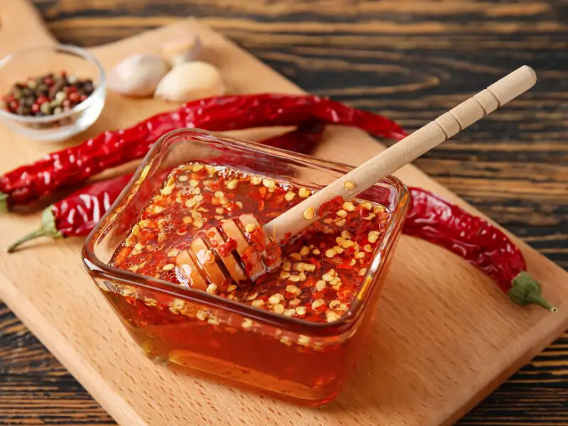 Spicy chili oil with red pepper flakes in a glass dish, garlic and dried chili peppers on wooden board